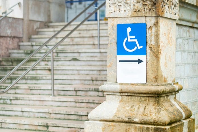 Addressing accessibility barriers in a Grade II listed building. Wheelchair accessibility sign mounted on a stone wall near outdoor stairs, representing inclusive design, accessibility, mobility rights, public infrastructure, and urban accessibility.