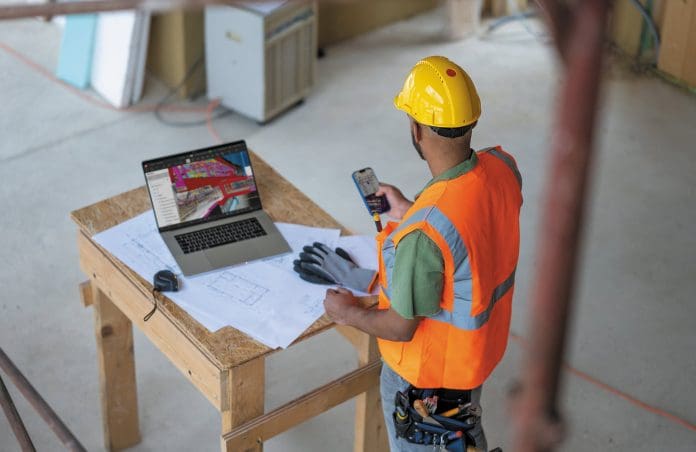 On-Site A worker in hi viz and yellow hard hat checking a phone. They are stood in front of a table covered with blueprints and a laptop