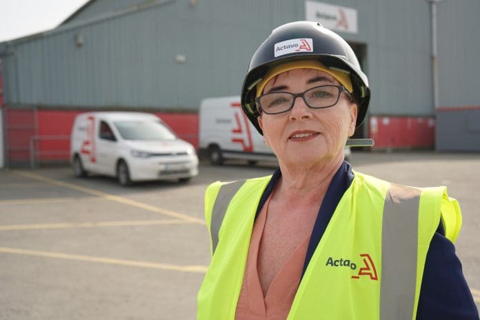 Karen Hendrick stood facing the camera in a yellow hi viz jacket and black hard hat in front of two vans and a warehouse