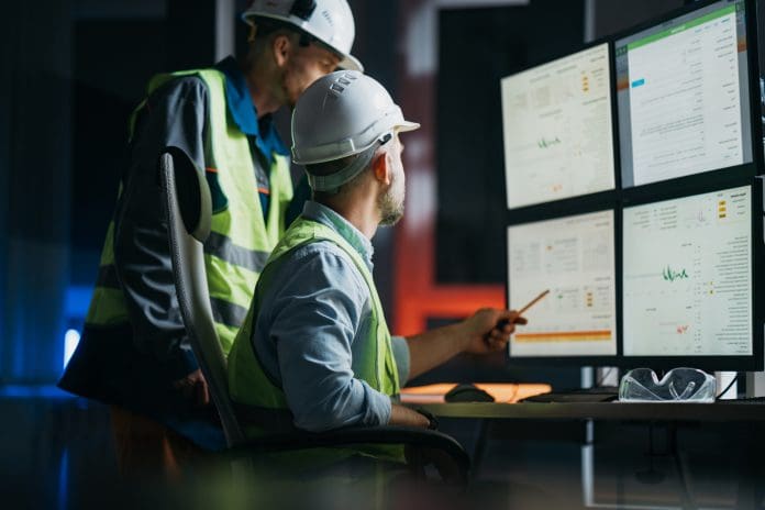 Group of operators control process on product line uses industry 4.0 and digital technology on modern factory. Two engineers follow assembly process uses SCADA system which AI technology and UI Poor product data costing UK construction £3.8bn a year