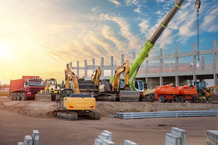 Many different multiclored colorful heavy industrial machinery equipment at construction site parking area against warehouse building city infrastructure development. Commercial vehicles rental sale Nationwide Hire files administration notice