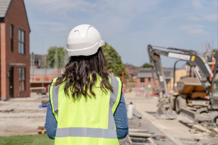 Woman builder engineer in bright safety jacket and hard hat uses surveying instrument at a construction site surrounded by construction materials and barriers Women in construction are key to solving the construction skills gap