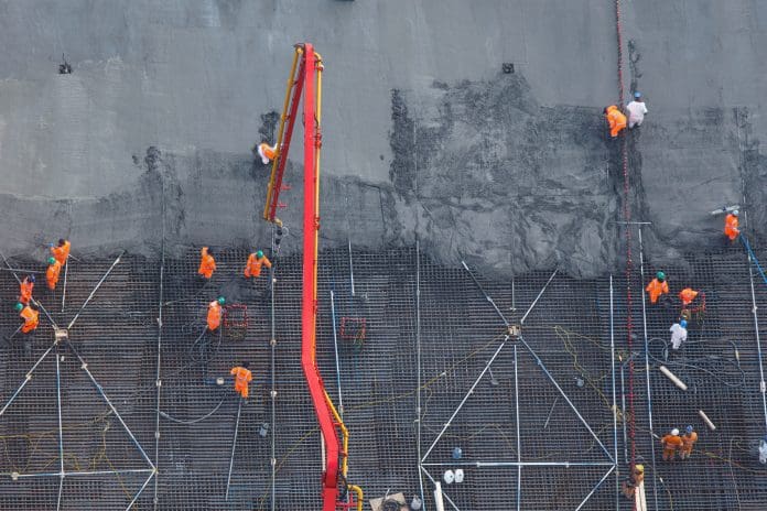 Aerial view of construction workers spreading fresh concrete from red pump truck boom across steel reinforced foundation grid at active commercial building site Overhaul of UK planning rules set to deliver fastest infrastructure in a generation