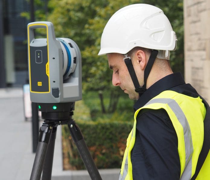 OLYMPUS DIGITAL CAMERA A male worker in hi viz and white hard hat standing next to a laser scanner