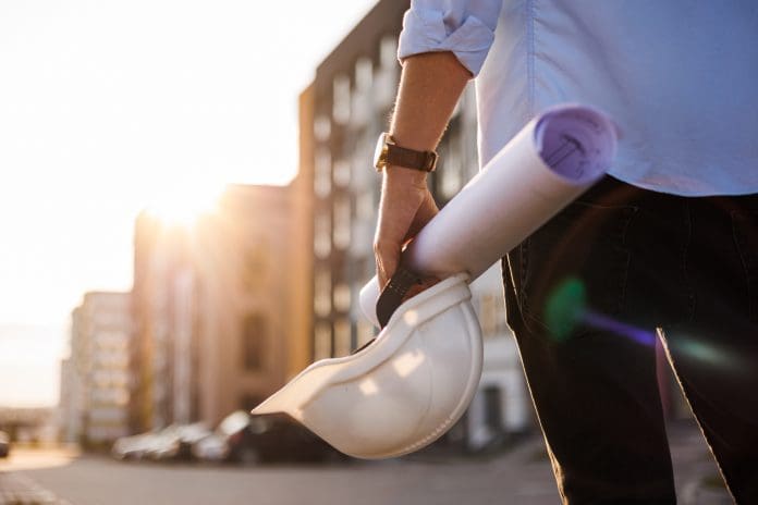 Unrecognisable engineer, architect or worker holds in hand protective white hard hat and blueprint on multi-apartment residential complex background, sunlighting. Concept of new real estate