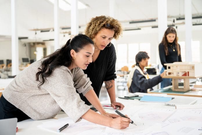 Teacher and student collaborating on architectural drawings with classmates in background Confidence increasing among architecture practices, RIBA finds