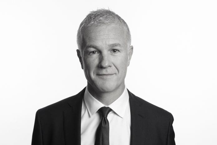 A black and white image of a man in a dark suit and tie with a white shirt stood against a white background