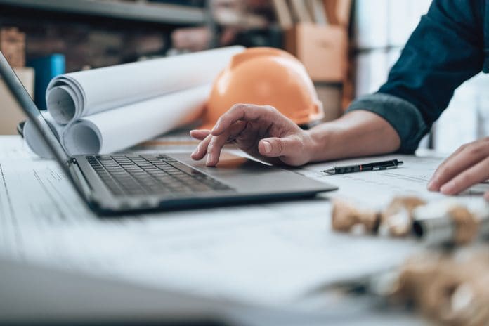 Engineering working with drawings inspection on laptop in the office and Calculator, triangle ruler, safety glasses, compass, vernier caliper on Blueprint. Engineer, Architect, Industry and factory concept. With construction administrations rising, cashflow resilience must become a priority