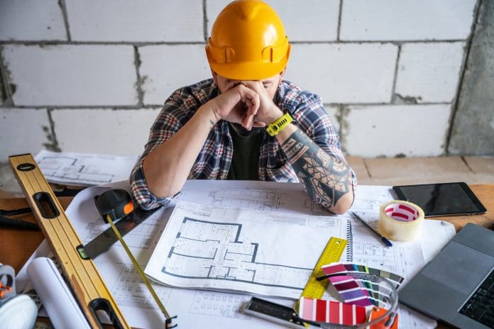 Tired construction worker at the table with a architectural blueprints and work tools The true cost of construction rework: Why getting it right first time matters