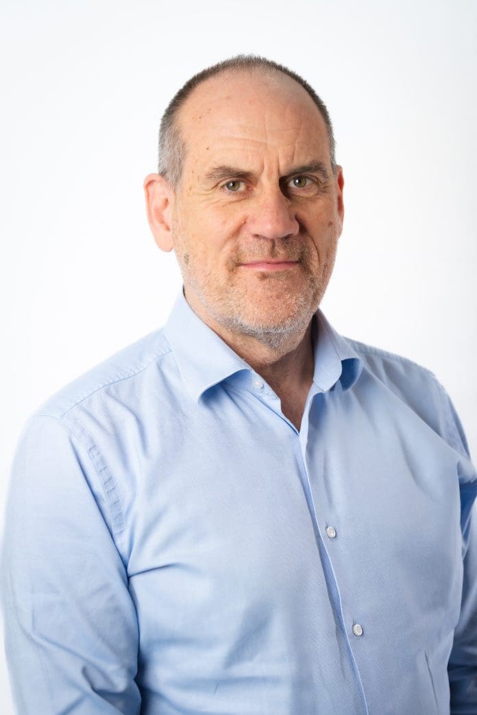 A man in a light blue shirt is stood against a plain white background looking towards the camera