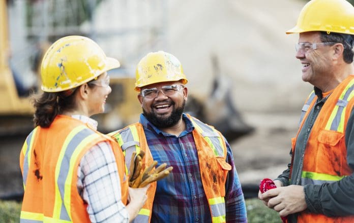 Three multi-ethnic construction workers chatting A group of three multi-ethnic workers at a construction site wearing hard hats, safety glasses and reflective clothing, smiling and conversing. The main focus is on the mixed race African-American and Pacific Islander man in the middle. The other two construction workers, including the woman, are Hispanic.