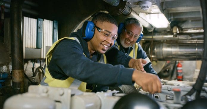 Senior expert mechanic coaching a trainee in a manufacturing plant. Smiling men in uniform working on engine repair in factory wearing safety glasses. Professional mechanic in development CITB report reveals regional disparities in construction apprenticeships