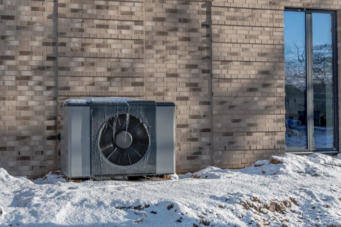 Air source heat pump unit with icicles hanging from casing installed against modern brick building surrounded by snow during winter. Energy-efficient residential heating technology. Heat pump handover: What to check before the first winter weather comes